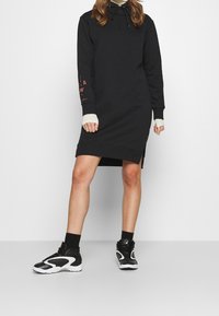 Černé mikinové šaty s kapucí, dlouhými rukávy, bočními rozparky a červeným textem na levém ruk ávu. V kombinaci s černobílými sportovními botami.