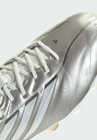 Chaussure de football Adidas argentée et blanche avec des lacets blancs et trois bandes blanches inclinées sur le côté, présentant une tige synthétique texturée.