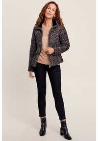 Gepolsterte schwarze Jacke mit einem strukturierten Zebra-Muster, ausgestattet mit einem Reißverschluss und hohem Kragen, getragen über einem hellbeigen Oberteil und schwarzen Skinny Jeans.