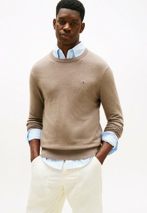 Suéter marrón de punto con cuello redondo, usado sobre una camisa de dress de rayas azul claro. Pantalones blancos de tela texturizada completan el conjunto.