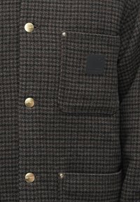 Abrigo de lana de cuadros marrones con botones dorados, un bolsillo en el pecho con una insignia negra y una superficie de tela texturizada.