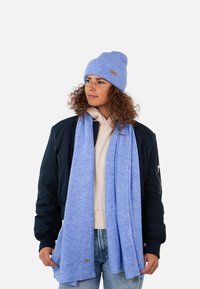 Bonnet et écharpe en tricot bleu clair fabriqués à partir d'un matériau doux, portés sur un hoodie crème et une veste bomber noire, avec un discret détail de logo.