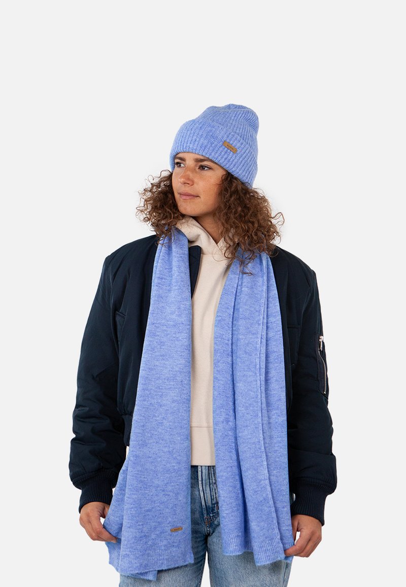 Bonnet et écharpe en tricot bleu clair fabriqués à partir d'un matériau doux, portés sur un hoodie crème et une veste bomber noire, avec un discret détail de logo.