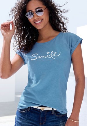 Himmelblaues T-Shirt mit hochgerollten Ärmeln, auf dem in geschwungenen weißen Buchstaben "Smile" steht. Kombiniert mit dunkelblauen Jeans und einem weißen Gürtel.