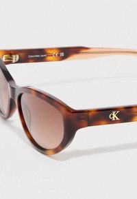 Okulary przeciwsłoneczne w kolorze żółwia z grubą, błyszczącą oprawką i brązowymi soczewkami z gradientem. Złote logo na zauszniki. Smukły, nowoczesny design.