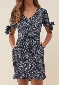 Granatowa sukienka w kwiaty z krótkimi rękawami, detalami w postaci wiązań, dekoltem w serek, talią z wiązaniem oraz widocznymi guzikami z przodu. Posiada boczne kieszenie.