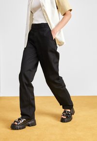 Black tailored pants with a straight cut, featuring pockets and a smooth fabric. Paired with black chunky sandals with studded accents.