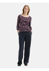 Blusa viola scuro con motivo floreale, maniche lunghe e texture leggermente arricciata; abbinata a pantaloni navy a gamba larga e calzature nere.