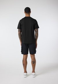 Black crew neck t-shirt, cotton material, relaxed fit. Paired with navy cargo shorts, two side pockets. White sneakers, visible tattoos on legs.