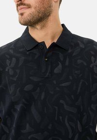 Marineblauwe polo met een textuurpatroon van abstracte vormen, een drievoudige knoopsluiting en een klein merkembleem op de borst.