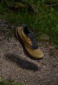 Des chaussures avec un design brun et vert olive, un dessus en mesh texturé, des accents noirs et une semelle rembourrée, suspendues au-dessus d'un chemin en gravier.