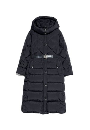 Femme portant un long manteau matelassé noir par-dessus un pull moutarde, un jean noir et des bottines noires, debout avec une main dans la poche.