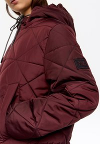 Bordowa pikowana kurtka z kapturem, z teksturowaną powierzchnią, z zapięciem na sznurek i czarną naszywką z logo na rękawie.