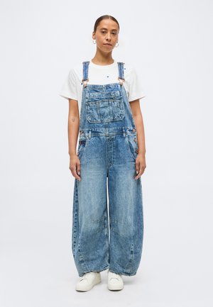 Femeie ținându-se în picioare, purtând salopetă din denim albastru deschis cu picioare largi, tricou alb, adidași albi, cu părul prins și cercuri aurii la urechi.