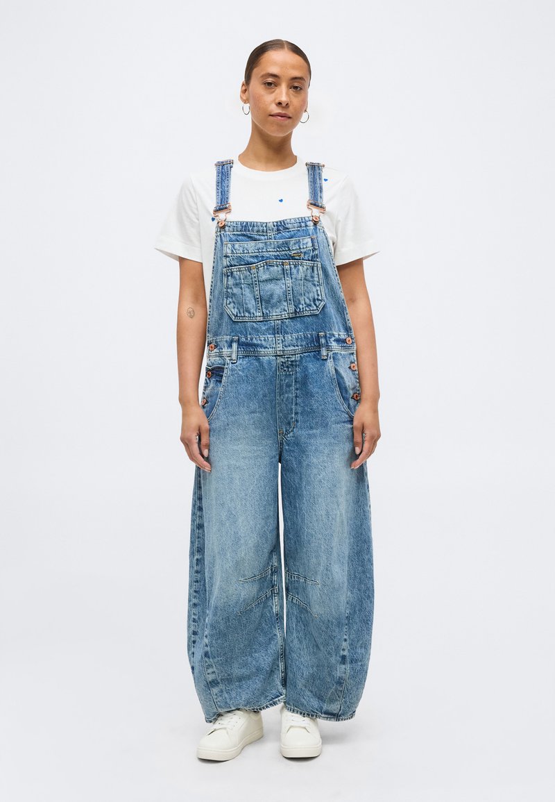 Γυναίκα που στέκεται, φορώντας φαρδιά ξεθωριασμένη μπλε τζιν σαλοπέτα, λευκό μπλουζάκι, λευκά αθλητικά παπούτσια, με μαλλιά δεμένα προς τα πίσω και κρίκους στα αυτιά.