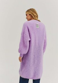 Dlhý levanduľový pletený kardigán s jemnou, nadýchanou textúrou, veľkými rukávmi a štítkom na chrbte. Nosený cez tmavomodré rifle.