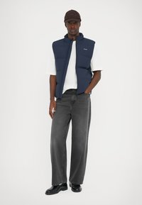 Vestă puffer de culoare navy peste o cămașă albă, asortată cu blugi negri largi și pantofi negri. Șapca este maronie cu un logo.