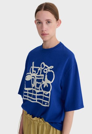 Kobieta o jasnobrązowych włosach, ubrana w niebieski, oversize'owy T-shirt z białym abstrakcyjnym wzorem naczyń kuchennych oraz musztardowe spodnie.