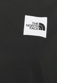 Černé tričko s bílým obdélníkovým znakem, na kterém je logo "THE NORTH FACE" v černém písmu a zakřivený design loga. Hladký povrch.