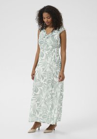 Robe maxi avec un motif fleuri vert menthe, décolleté volanté, taille cintrée et jupe fluide. Portée avec des sandales à talons beiges.