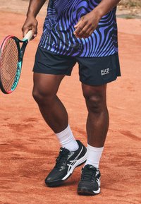 Pantalones cortos atléticos negros con el logo de EA7, combinados con zapatillas de tenis en blanco y negro y calcetines tobilleros blancos, sosteniendo una raqueta de tenis.