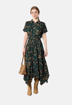 Woman wearing a green floral midi dress with short sleeves, holding a tan suede handbag, and wearing knee-high tan boots.