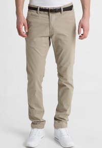Beige Chinos aus Baumwolle in schmaler Passform. Mit einem Reißverschluss, fünf Taschen und einem braunen Leder-Gürtel ausgestattet. Werden mit weißen Sneakers getragen.