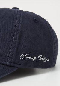 Tamsiai mėlynas medvilninis beisbolo kepuraitė su lenktu kraštu ir balta siuvinėta "Tommy Hilfiger" logotipu šone. Matomas tekstūruotas audinio detalė.