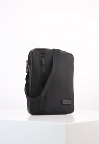 Borsa a tracolla nera Calvin Klein con tasca frontale con zip e tracolla regolabile su superficie bianca.