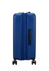 Valise rigide bleue avec des rainures verticales, fermeture éclair, poignée latérale noire et quatre roues pivotantes. Surface en plastique lisse et durable.
