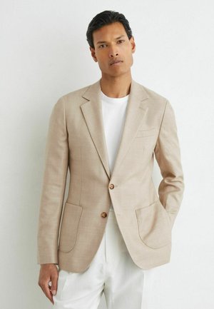 Mand iført en beige blazer over en hvid skjorte og hvide bukser står foran en hvid baggrund med den ene hånd i lommen.