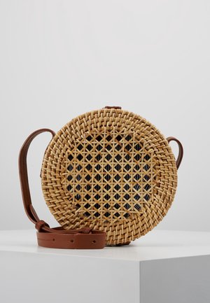 Sac bandoulière rond en rotin tressé avec motif géométrique et bandoulière en cuir marron réglable sur surface blanche.