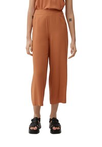 Pantalon court ample de couleur rouille avec une texture lisse, doté de poches latérales et d'une taille haute. Porté avec des sandales compensées noires.
