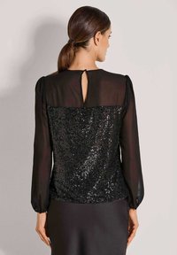 Schwarzes Top mit einem paillettenbesetzten Rückenteil, durchsichtigen langen Ärmeln und einem Schlüssellochdetail am Hals. Glatte Textur mit einer figurbetonten Silhouette.
