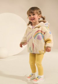 Veste en polaire couleur crème avec des motifs arc-en-ciel, associée à des leggings jaunes ornés d'accents étoilés et des baskets multicolores.