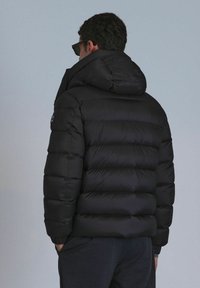 Black puffer jacket with quilted design, hood, and elastic cuffs. Smooth texture and compact shape, featuring a small logo on the sleeve.