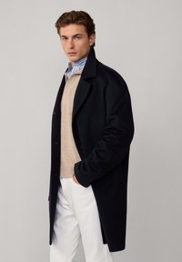 Cappotto di lana navy con un design su misura, indossato sopra un maglione beige e una camicia colletto azzurro chiaro, abbinato a pantaloni bianchi.