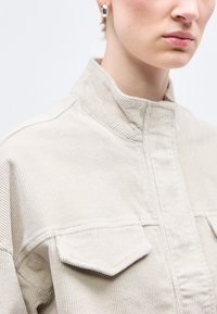 Κοντινό πλάνο ενός ατόμου που φορά μια μπεζ ζακέτα κοτλέ με εμπρόσθιες τσέπες με καπάκι και ασημένια σκουλαρίκια κρίκους, με απλό φόντο.