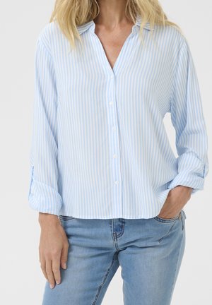 Frau trägt ein hellblau-weiß gestreiftes Hemd mit Knöpfen und hochgekrempelten Ärmeln sowie hellblaue Jeans, eine Hand in der Tasche.