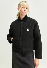Schwarze Jacke mit Reißverschluss vorne, samtigem Kragen und Fronttaschen. Verfügt über ein kleines weißes Logo-Patch und ein leicht verkürztes Design.