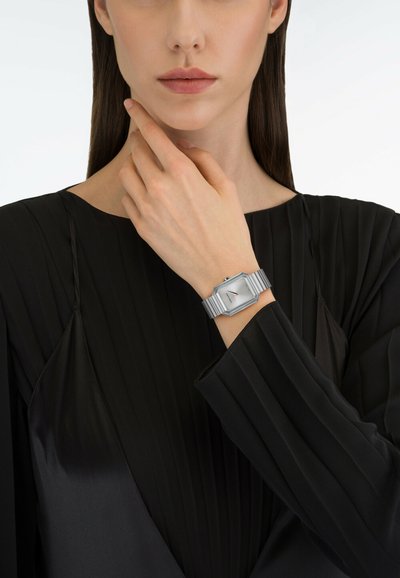 Hõbedane kuusnurkne käekell hõbedase rihmaga, asetatud käele, mis puhkab näo lähedal, lihtsa valge tausta ees.