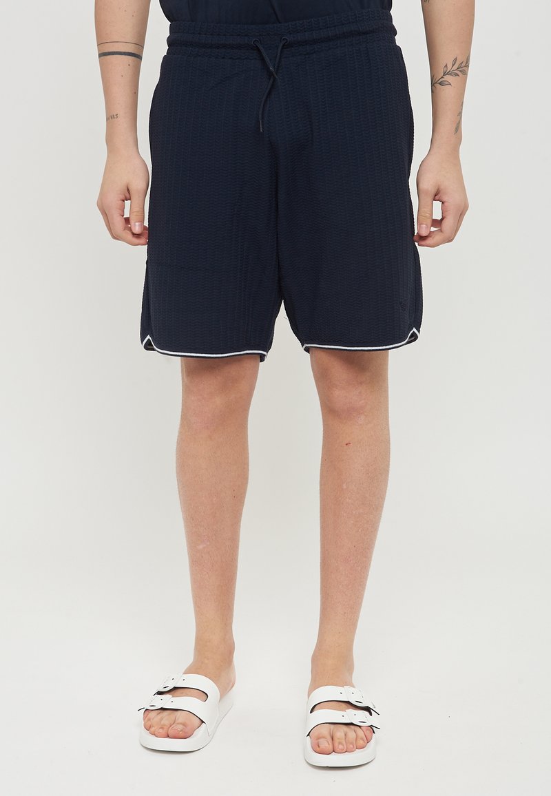 Pantaloni corti blu navy con un motivo testurizzato, vita elastica e stringa, con bordi laterali bianchi e lunghezza moderata. Indossati con sandali bianchi.