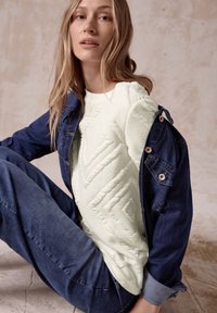 Cremfarbener Pullover mit geometrischen Mustern, unter einer blauen Jeansjacke getragen. Das Model sitzt poseierend in blauen Jeans vor einem neutralen Hintergrund.