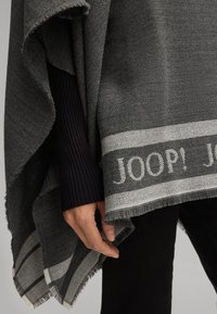 Poncho grigio con orlo sfilacciato, caratterizzato da un motivo a righe e la parola "JOOP!" in bianco. Indossato sopra una maglia nera a coste.