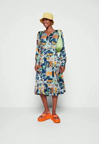 Mulher vestindo um vestido de patchwork floral colorido, chapéu de balde amarelo, bolsa verde de ombro e sandálias de plataforma laranja brilhante, sorrindo.
