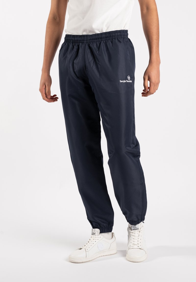 Navy blue, lightweight track pants with elastic waistband and cuffs. Features white logo details. Paired with white sneakers.