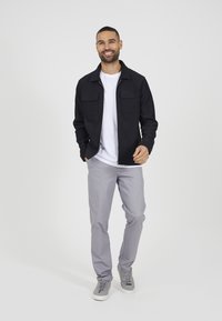 Schwarze gerippte Zip-Jacke mit zwei Vorderaschen, getragen über einem weißen T-Shirt, kombiniert mit hellgrauen Hosen und grauen Turnschuhen.