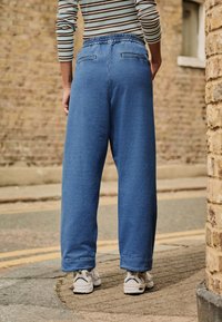 Blugi evazați albaștri cu talie elastică, prevăzuți cu două buzunare în spate și tivuri răsucite, purtați cu adidași albi pe o stradă pietruită.