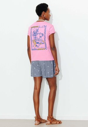 Femme portant un t-shirt rose avec une illustration et un texte sur le petit-déjeuner, un short bleu à carreaux, des sandales marron et un collier blanc, debout de profil.