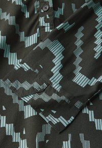 Camicia nera con un motivo geometrico turchese, dotata di chiusura a bottoni e polsini ripiegati. Il materiale sembra liscio e ha una leggera lucentezza.